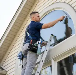 Professional window cleaner on a ladder using a squeegee to clean an exterior arched window, Louisville, KY.